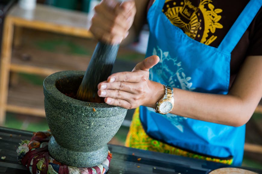A woman in a light blue apron, whose head we cannot see, is using a gray pestle and mortar to grind herbs as part of a cooking class in Bangkok.