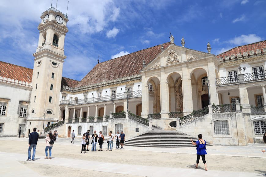 Människor tar foton framför Coimbra University, en enorm vit byggnad med ett tillhörande torg.  Byggnaden har också flera stora pelare nära ingången och ett högt klocktorn.