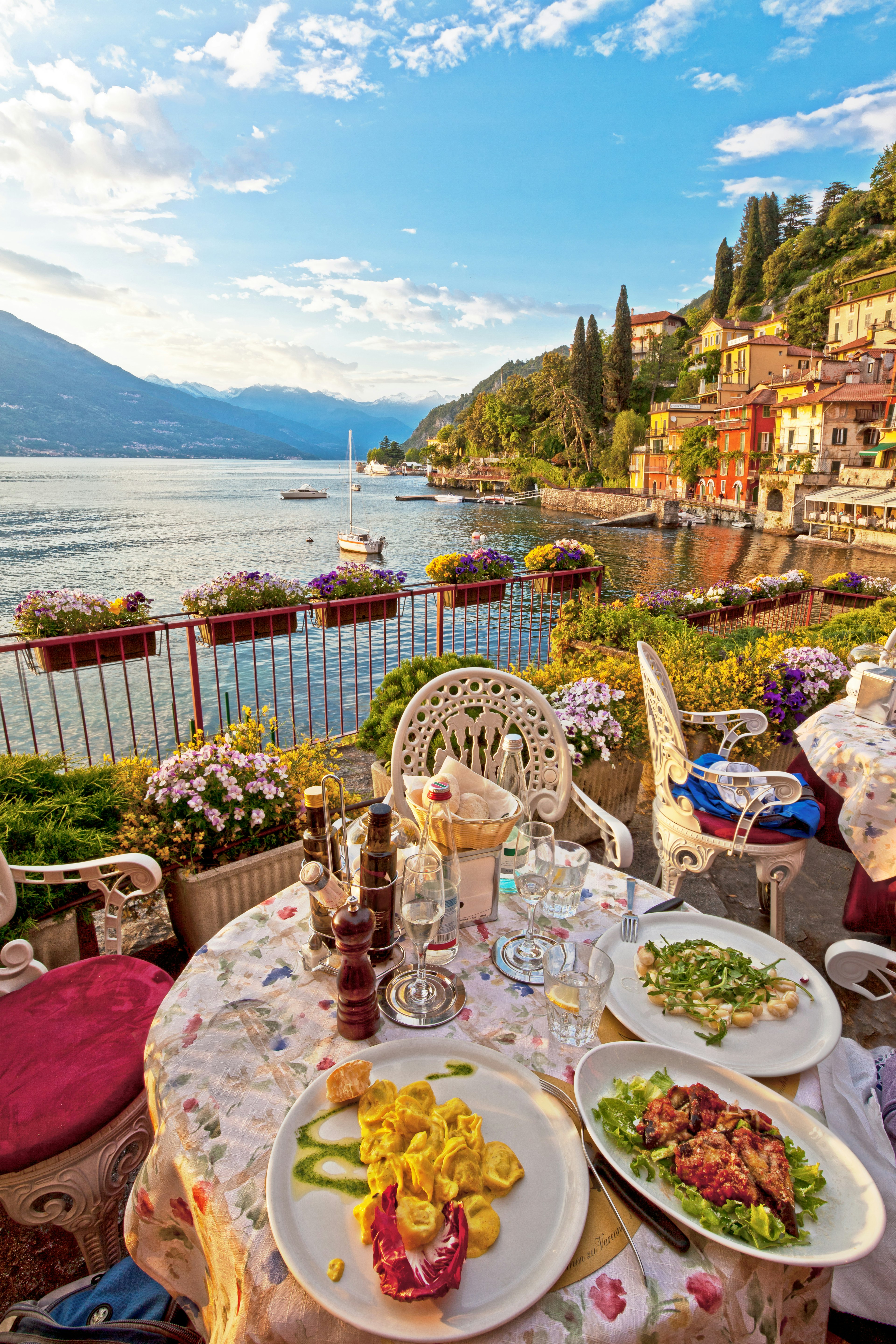 Three plates of lovely vegetarian Italian food on white plates on a table on a lovely outdoor terrace overlooking a beautiful, calm lake, with antique buildings and alpine mountains in the background