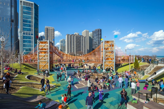 Adults and children in a large playground, with a big wooden climbing structure and metal tube slides