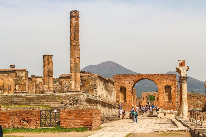 Ruinstaden Pompeji.  Människor går runt ruinerna av den tidigare staden, som förstördes av Vesuvius.  Vulkanen syns i bakgrunden av bilden. 