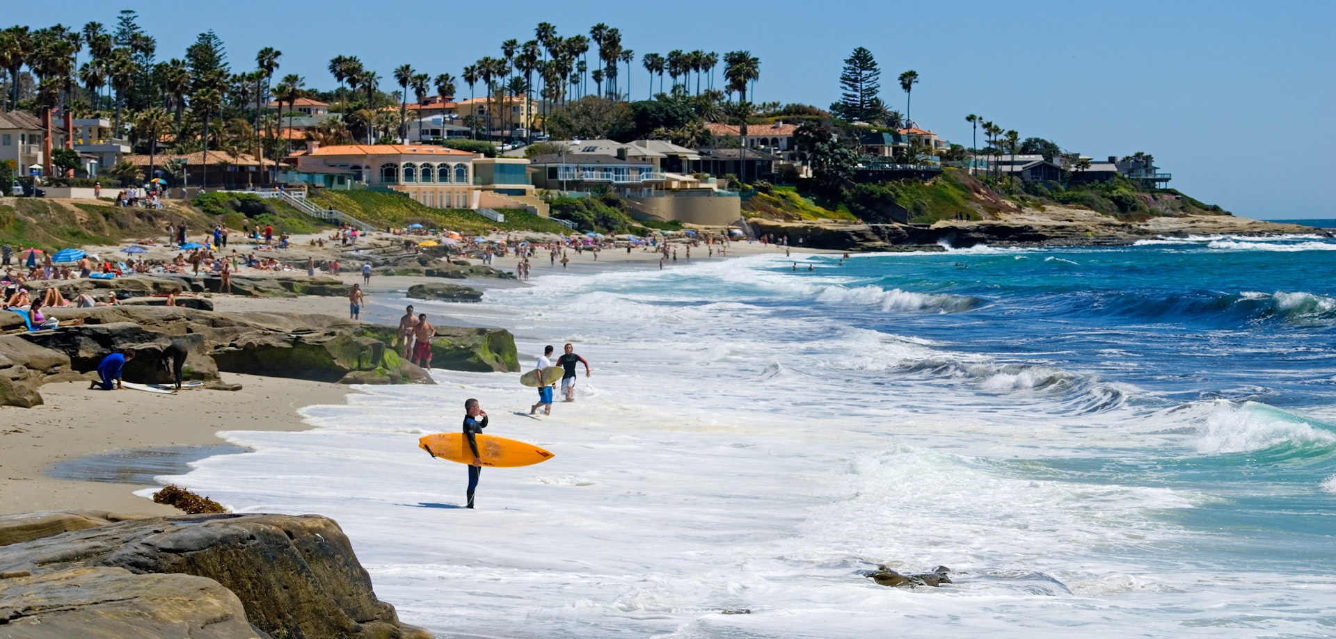 A surfer holding an orange board surveys the waves from the shore. The beach is busy with people.