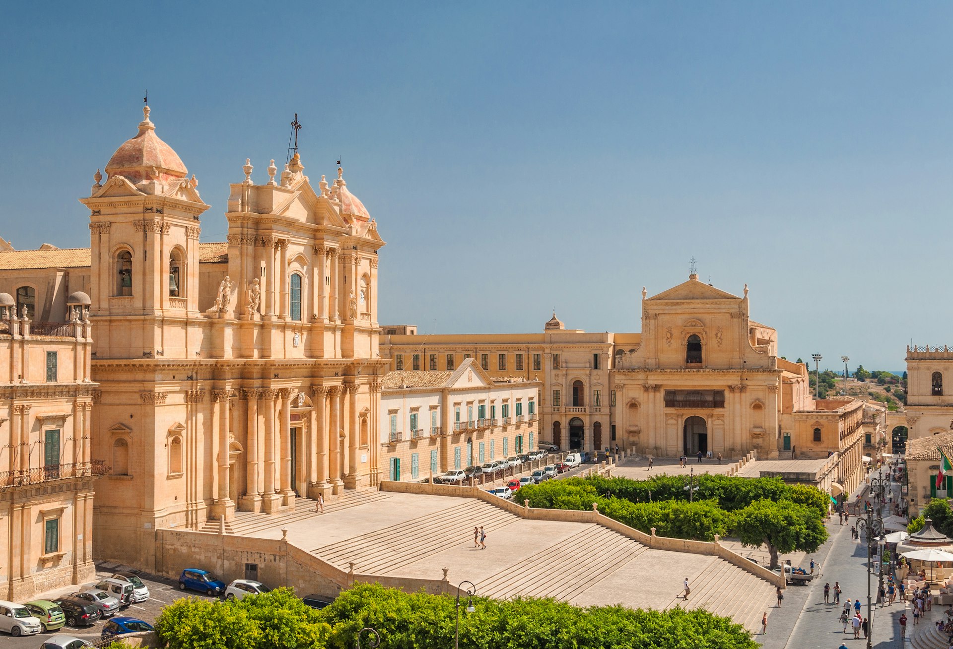 An aerial view of Noto Cathedral in the town of Noto, Sicily. The cathedral is grand, built from white limestone, and has a large flight of stairs leading up to it.