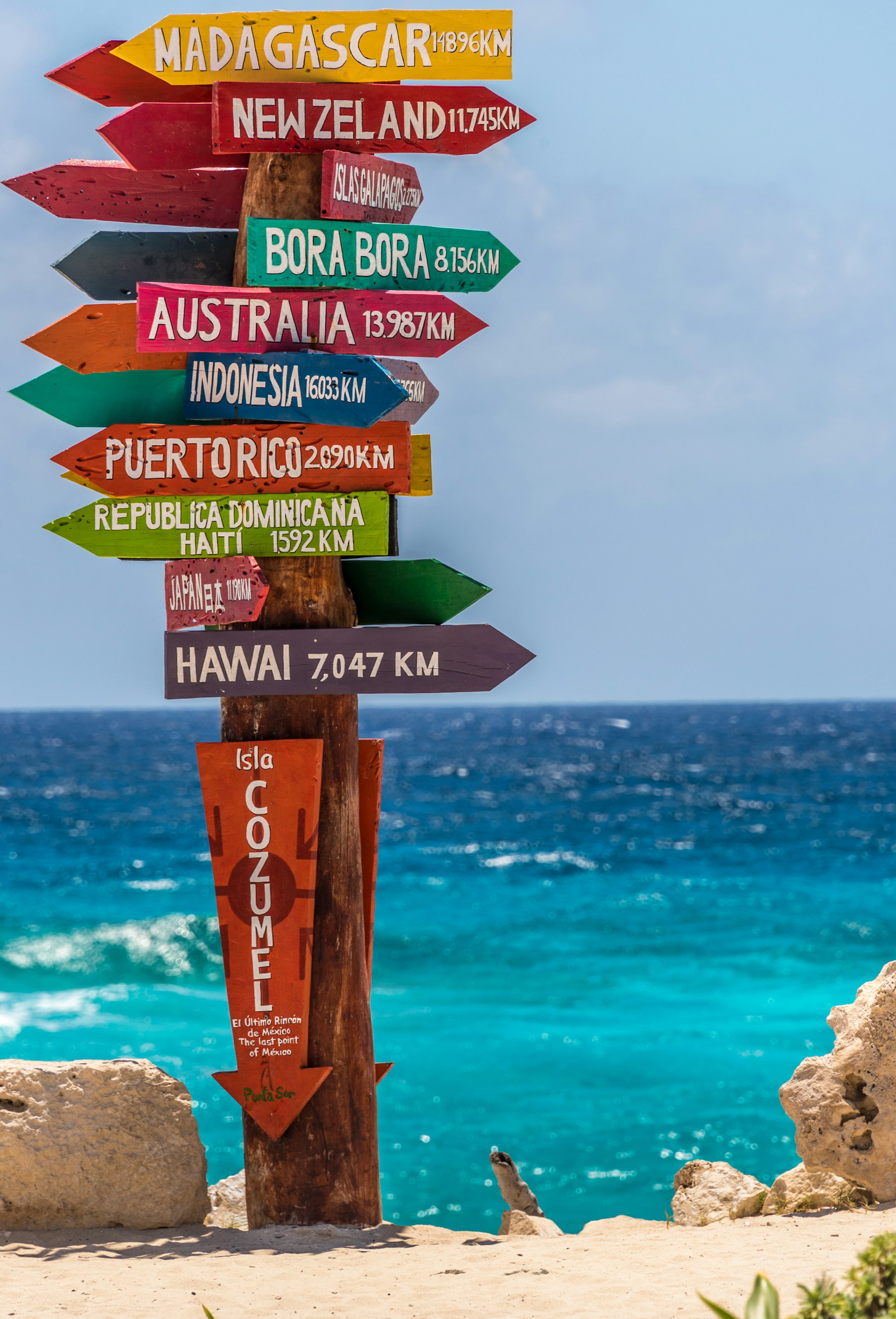 A colorful signpost near a beach shows many different arrows and distances to different locations worldwide