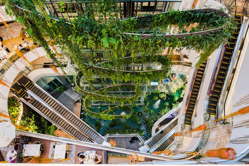 The Helix Quarter at Emquartier Shopping Mall in Thailand as seen from the top floor, with escalators, shops, and green interior plants for decoration all on view.