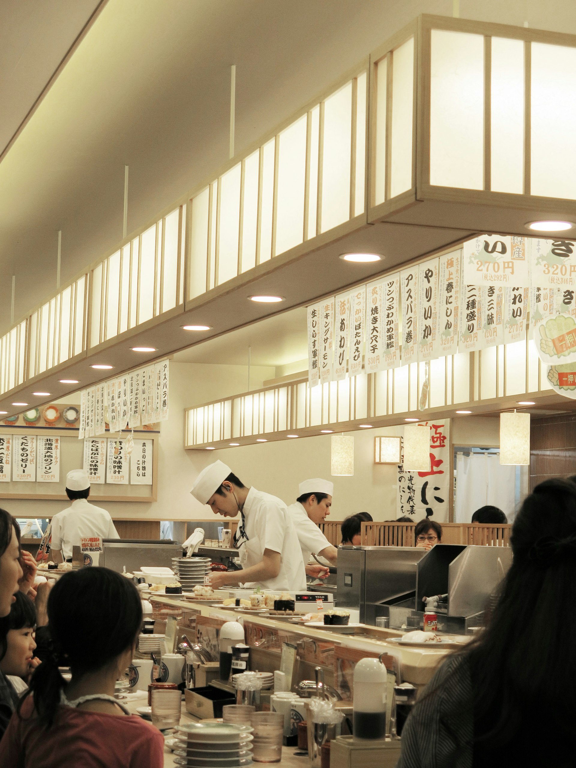 Chefs in white clothes with chef hats work away in a central kitchen. Diners sit at counters around the outside