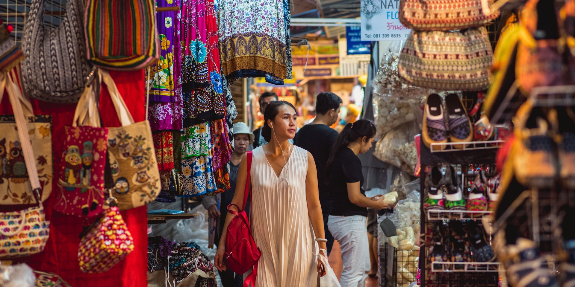 Young Asian woman walks with headphones along stalls and stands of Chatuchak market. 