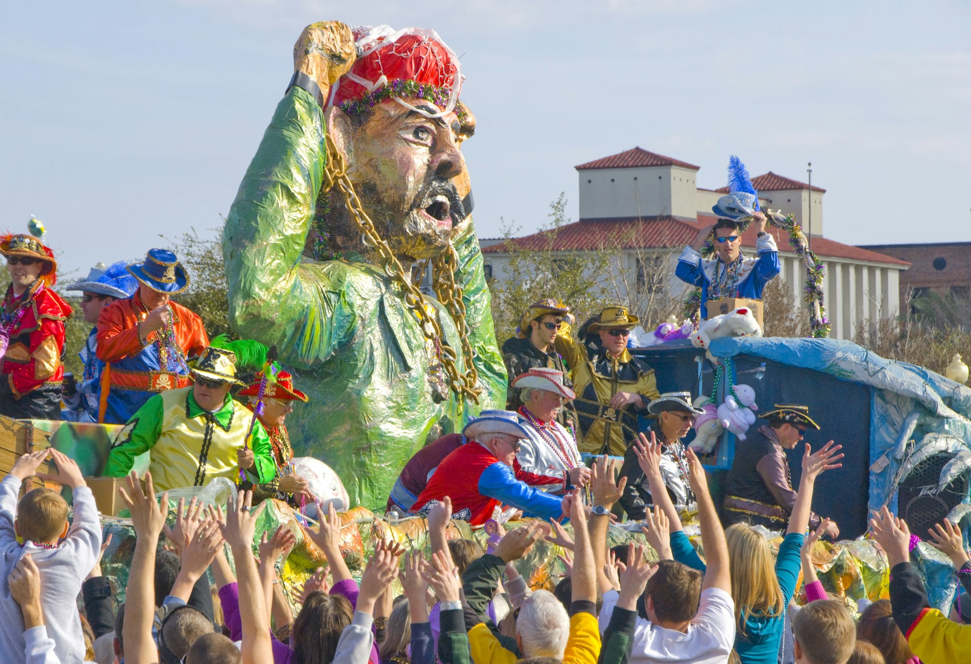 A float in a carnival has a large papier mache figure. People on the float are handing out mardi gras beads to revellers 