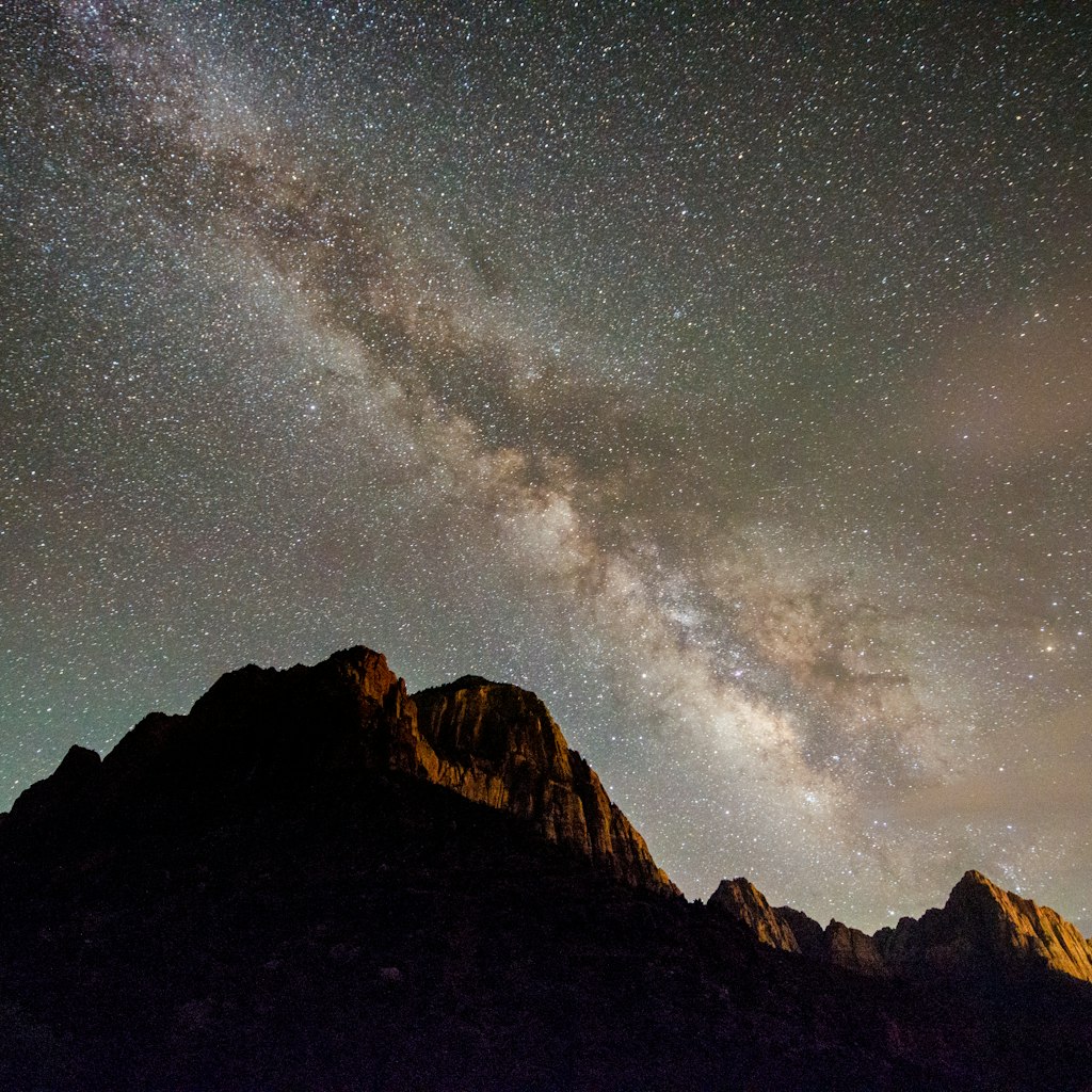 This is a horizontal, color photograph of the Watchman with the Milky Way at night in the Rocky Mountain landscape of Zion National Park in Utah, USA  in springtime. Light pollution illuminates the mountains.