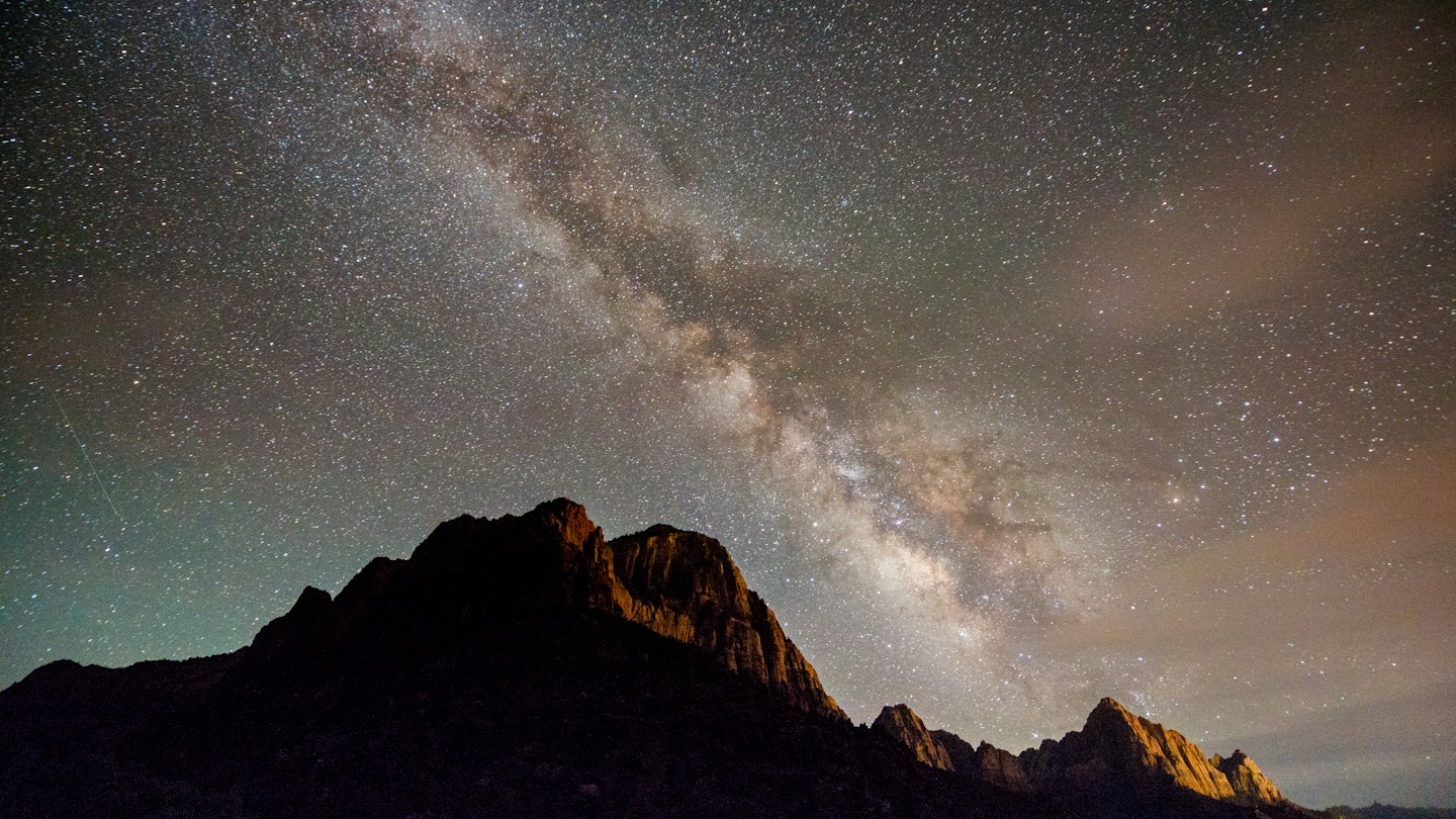 This is a horizontal, color photograph of the Watchman with the Milky Way at night in the Rocky Mountain landscape of Zion National Park in Utah, USA  in springtime. Light pollution illuminates the mountains.