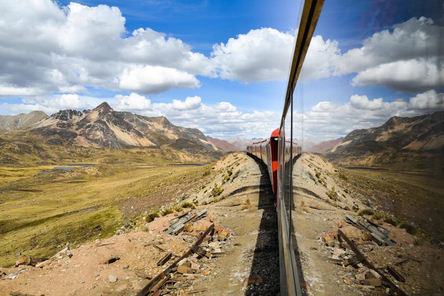 A side on view of a train travelling across the Peruvian landscape. The image is taken by someone leaning out of the moving train, with the Andes mountains reflected in the windows of the train.