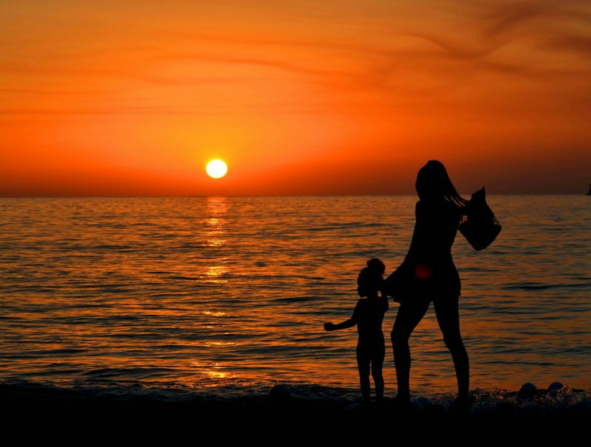 A woman and a young child are in silhouette as they walk on the beach at sunrise