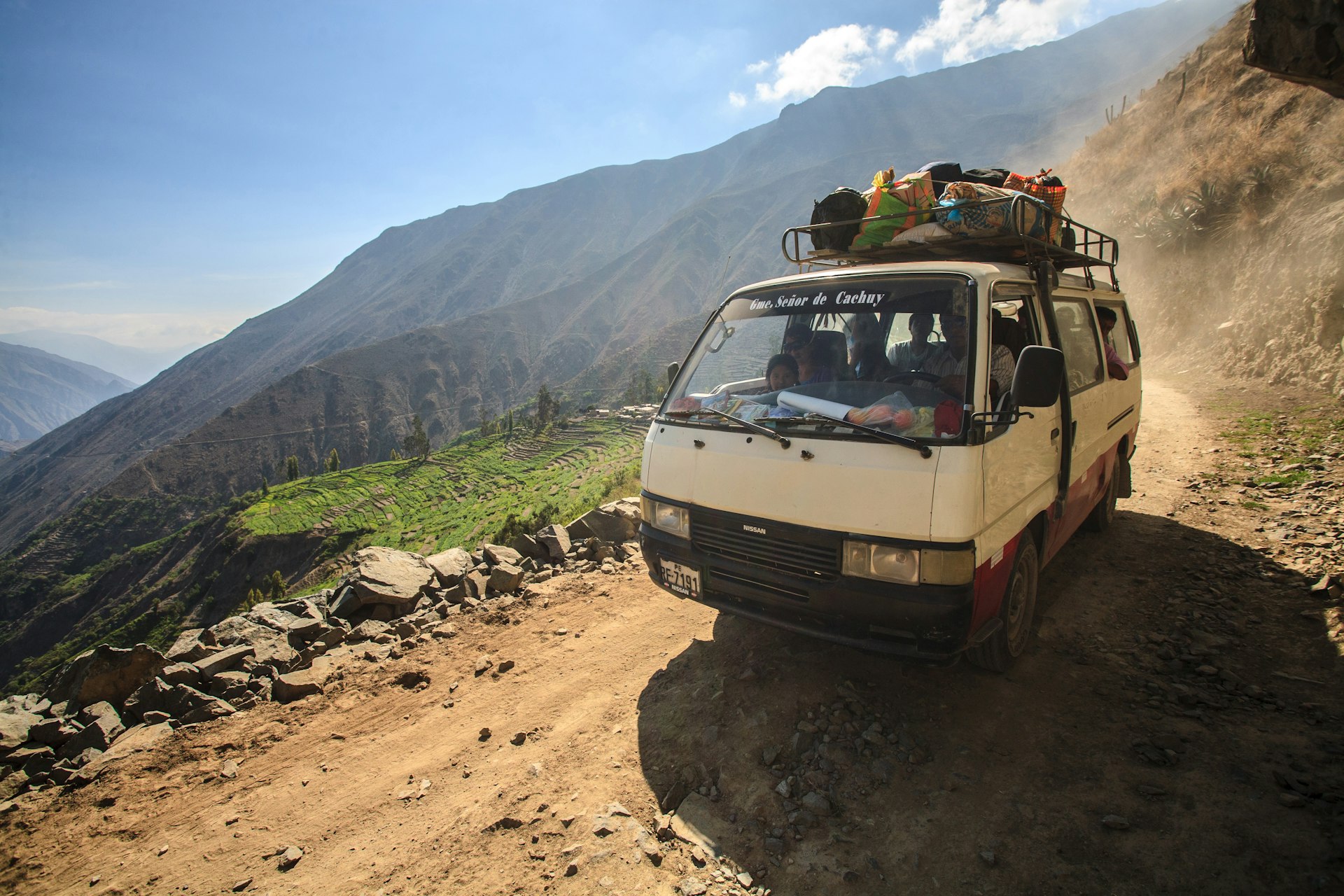A small, old bus drives along a mountain dirt road in the Peruvian Andes mountain range. Next to the road is a steep drop leading down into a green valley.
