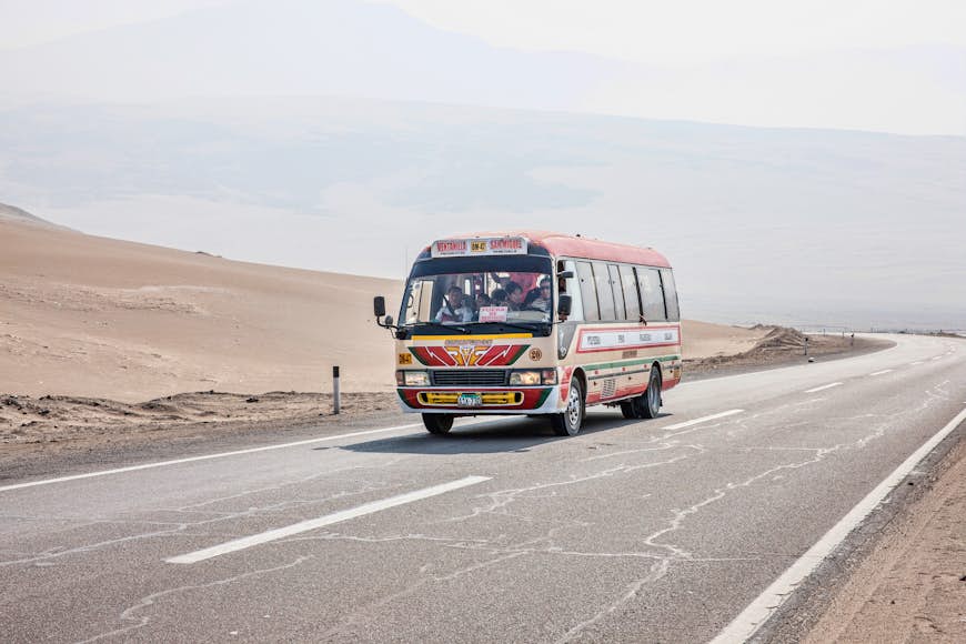A bus travels on the Pan-America Highway through Peru. The bus is painted in bright colours and a few of the passengers are visible through the windows. The landscape around the bus is desert.