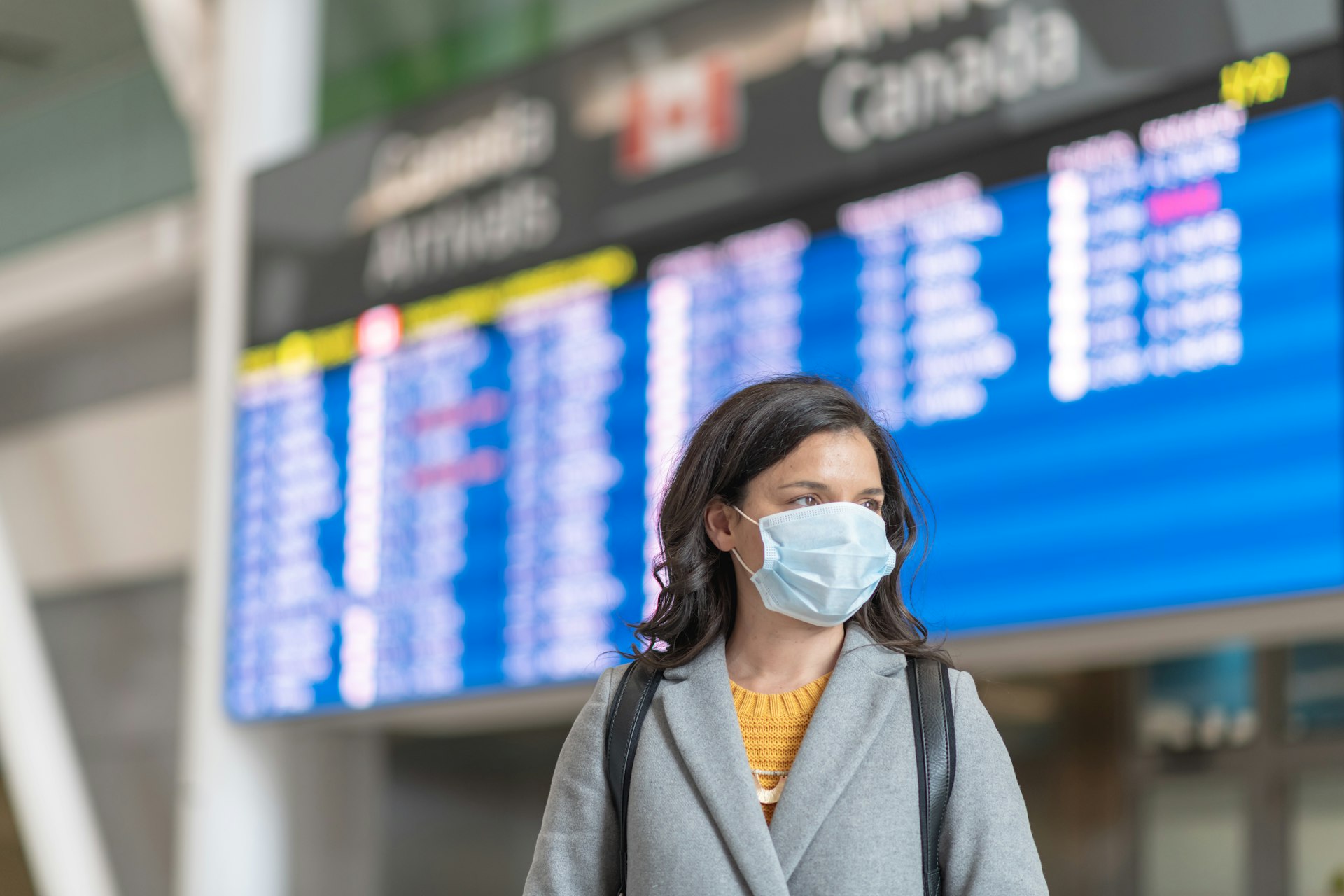 A solo travellers flight has been cancelled. She is standing in front of the departures board. She is wearing a protective face mask