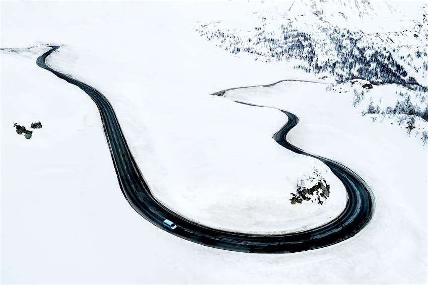 Blick aus der Vogelperspektive auf eine schwarze Bergstraße, die sich auf dem Weg nach St. Moritz durch eine verschneite Schweizer Landschaft schlängelt.  Ein einsames Auto fährt die Straße entlang, die im Angesicht des Schnees steht.