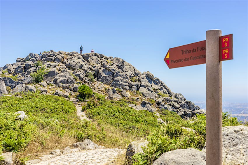 Uma placa aponta na direcção de Foia, o ponto mais alto do Algarve, em Portugal.  Um andarilho está no topo do cume ao fundo.
