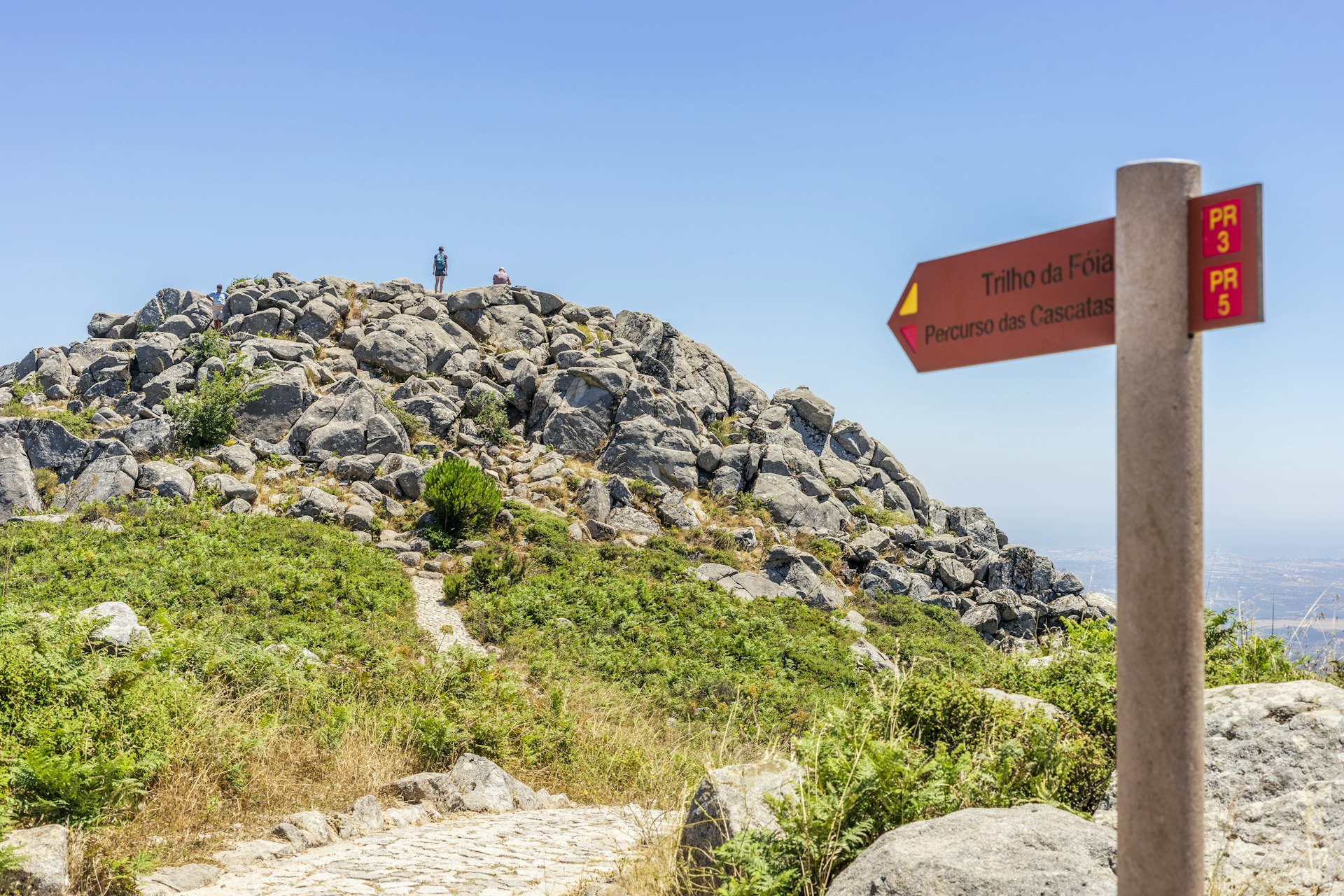 A sign points in the direction of Fóia, the highest point of the Algarve in Portugal. A hiker stands on top of the peak in the background.