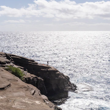 There is a fisherman about to cast his line in the water over a tall cliff. Spitting Caves Hawaii is a hidden gem of East Oahu.