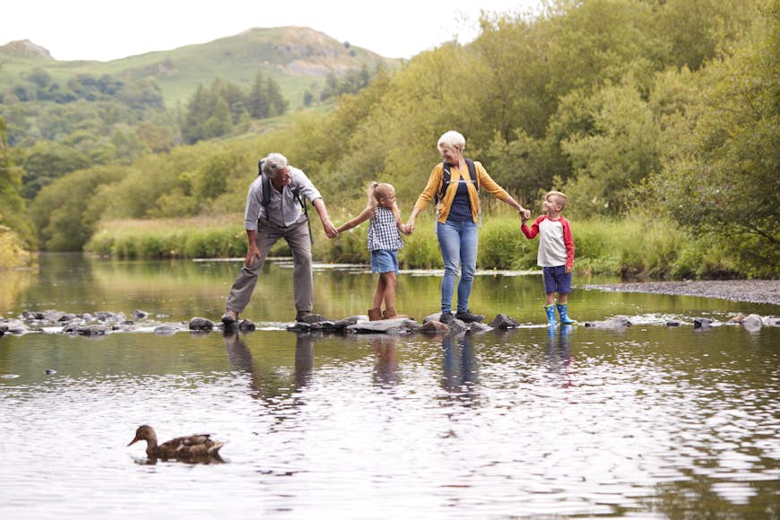 A family group of two adults and two children hold hands on a set of stepping stones in a lake.  There is a duck in the foreground and rolling green hills behind.