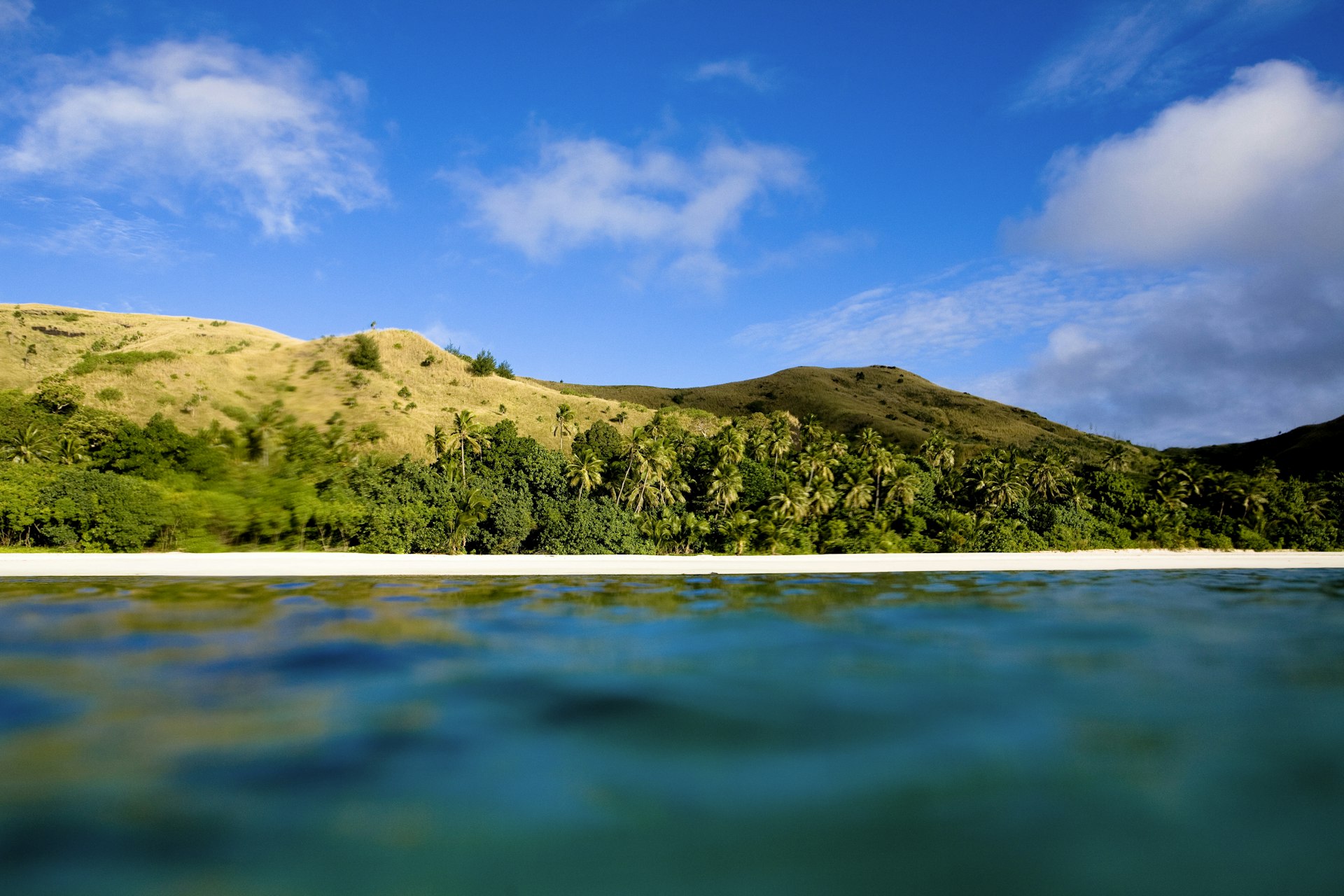 A view of the small island of Nacula, Fiji, from the water. The camera is just above the water level, meaning only a section of the white beach, lush forest and rolling hills of the island are visible.