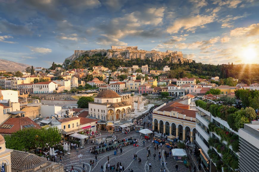 A high angle view of the cityscape of Athens. In the foreground a public square is bustling with people, while in the background a temple is visible on a high hilltop. Between the two, narrow laneways run between buildings and houses.