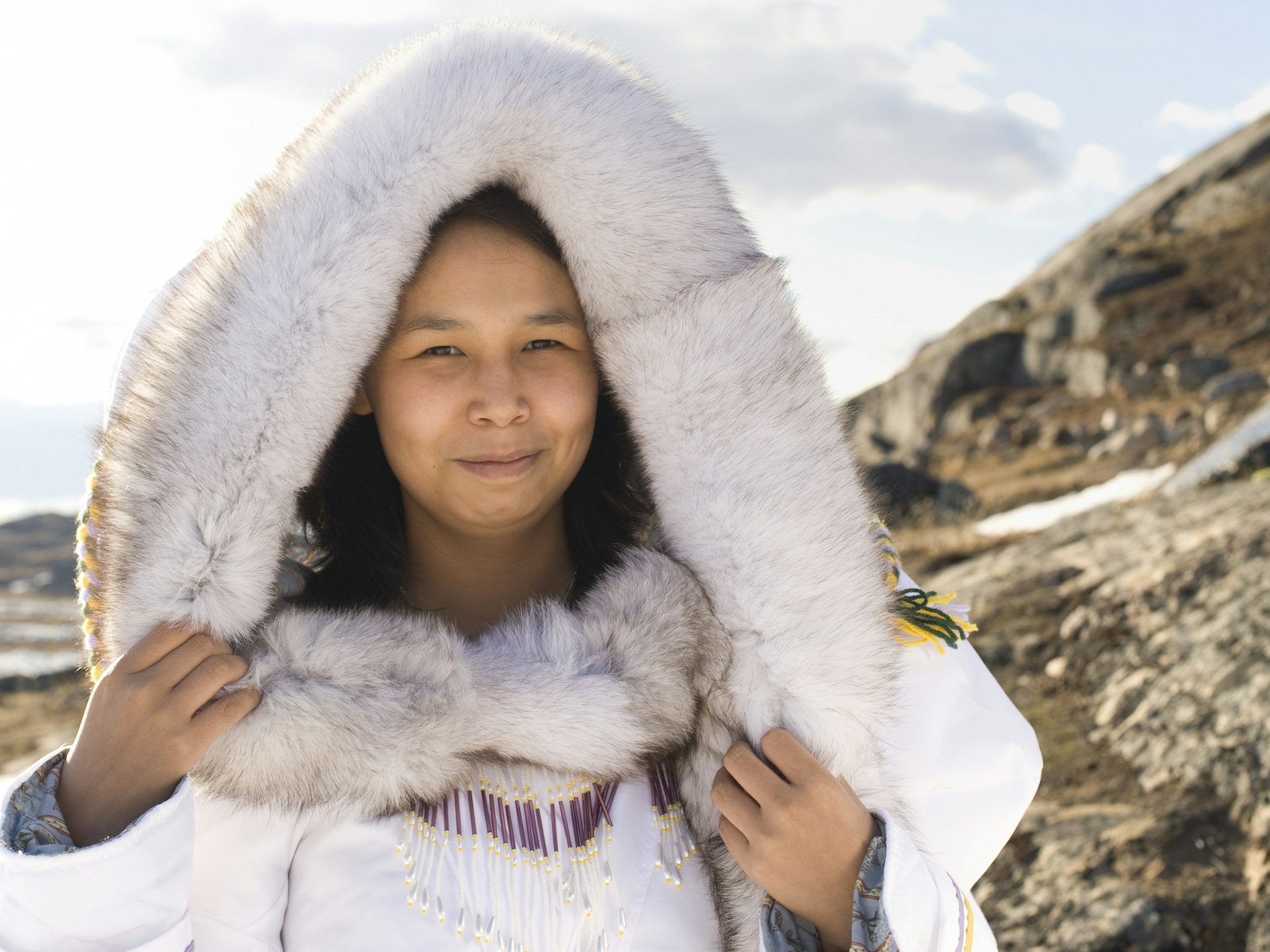 Picture of an Inuit woman standing on a tundra in Baffin Island, Canada. The woman is wearing a thick fur hood over thick white traditional clothing. The woman is wearing a necklace decorated in white and purple. The sky above the tundra is filled with cotton white clouds.
