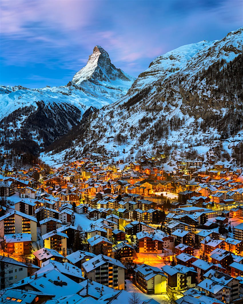 Luftbild des Zermatter Tals in Richtung Matterhorn.  Die Stadt hat viele Gebäude, in denen Lichter in den Fenstern sichtbar sind.  Der Gipfel des Matterhorns ist zerklüftet und mit Schnee bedeckt.