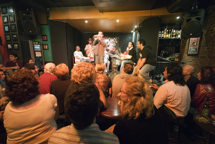 A flamenco jam session takes place at Cardamomo in Madrid.  A crowd of people sit at tables while musicians perform on a stage in the background of the image.