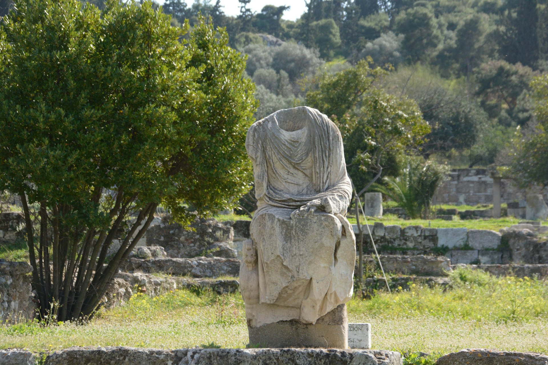 A headless Greek statue on display at the Ancient Agora archaeological site in Athens, Greece with olive trees in the background