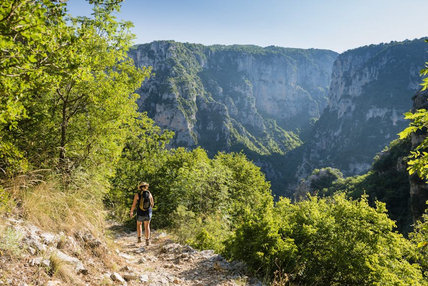 A hiker walks along a cliffside path into Vikos Gorge. The large valley gorge looms large in the background, with rocky vertical walls on either side.