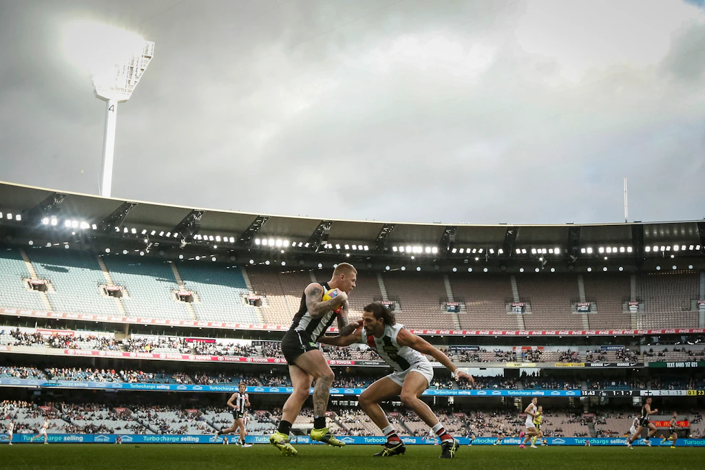 Two players tussle on the pitch of Melbourne Cricket ground during an AFL match between Collingwood Magpies and St Kilda Saints. The large grandstand of the stadium is visible in the background, with many fans watching on.