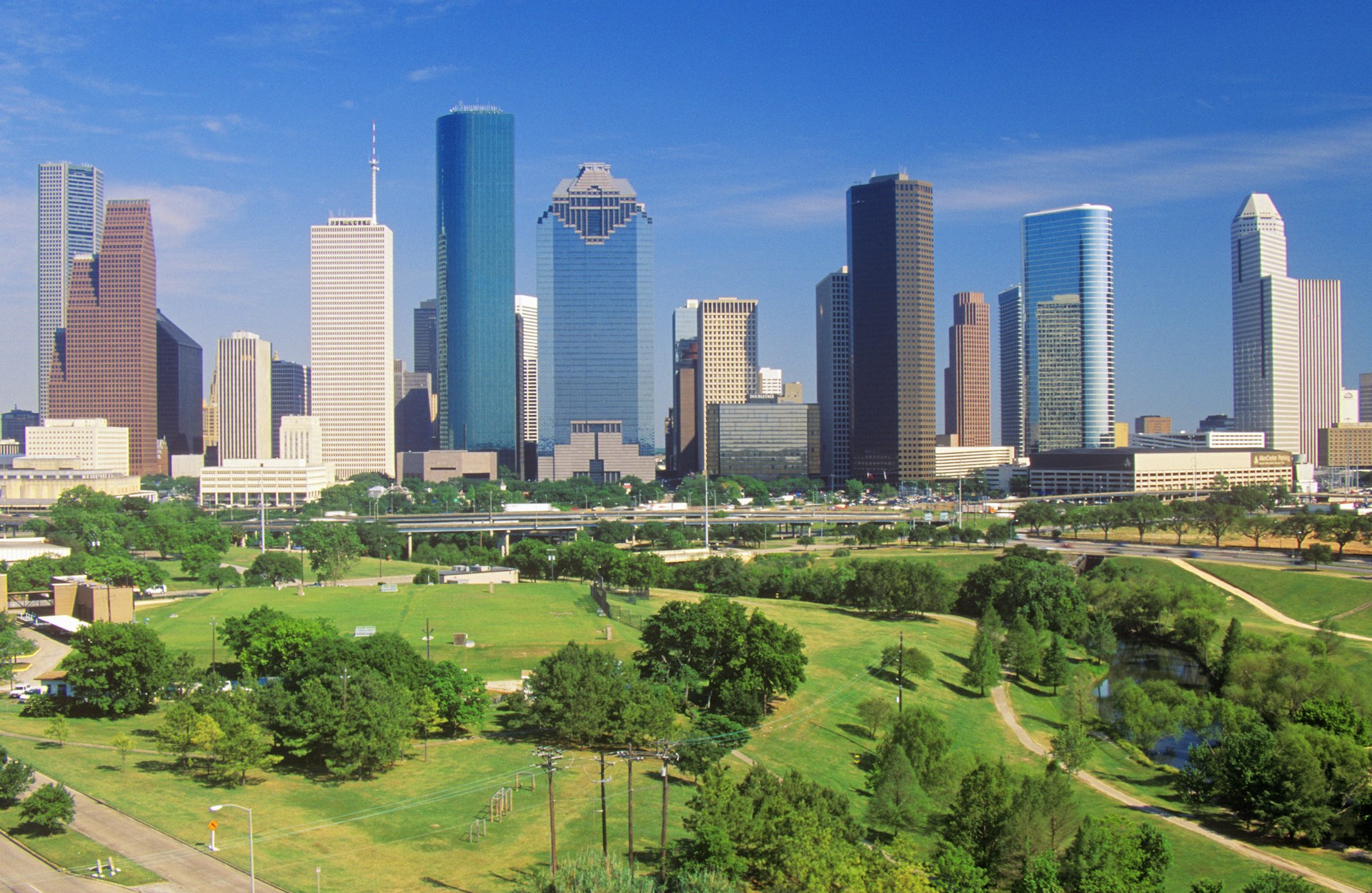A distant view of Houston's skyline in the afternoon with Memorial Park in the foreground. The park is an expansive carpet of green, dotted with trees.