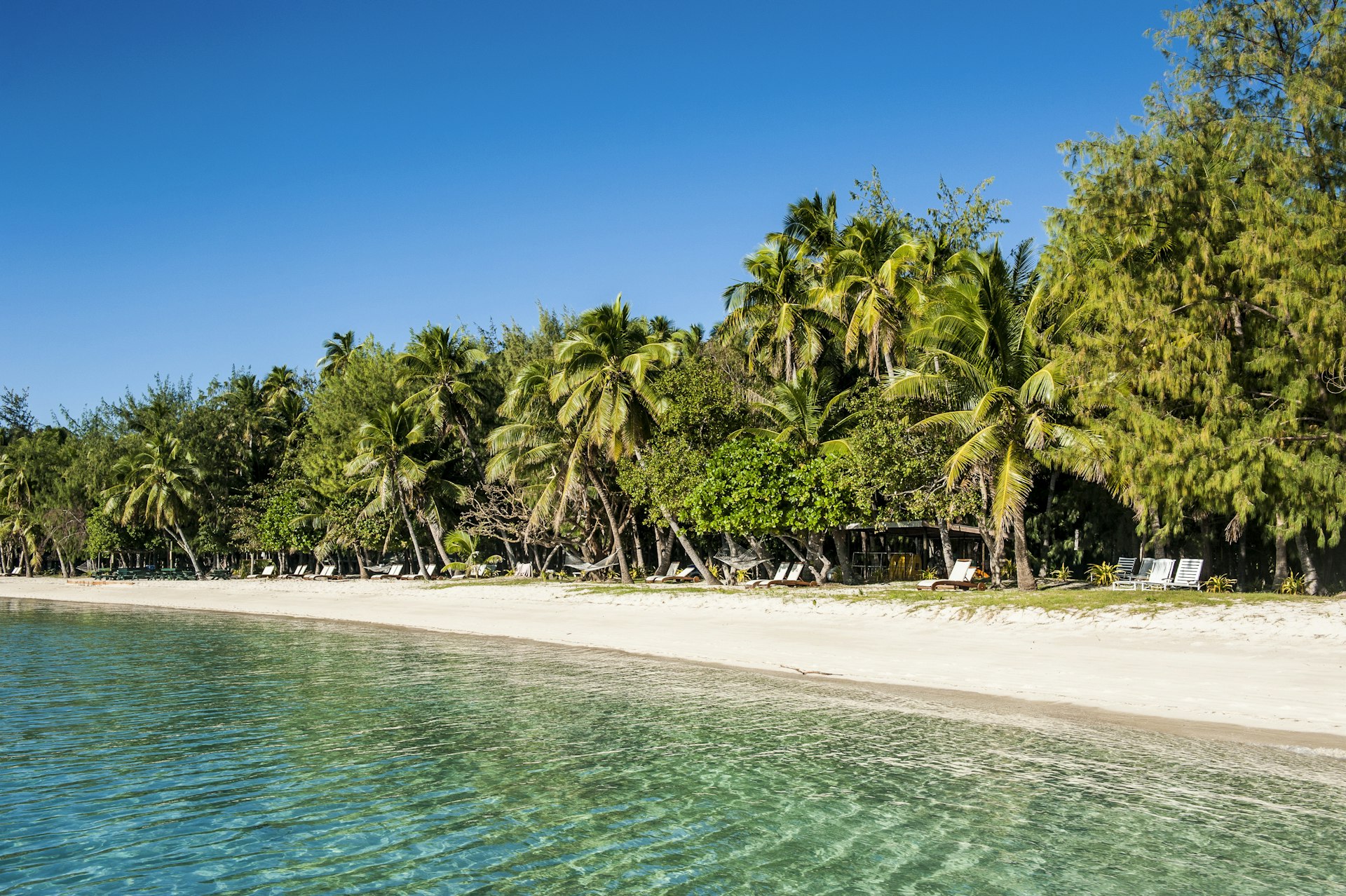A view of a white sand beach from the water. The waters lapping the beach are still and turquoise, while the forest behind it is dense and green. A blue sky is visible overhead.