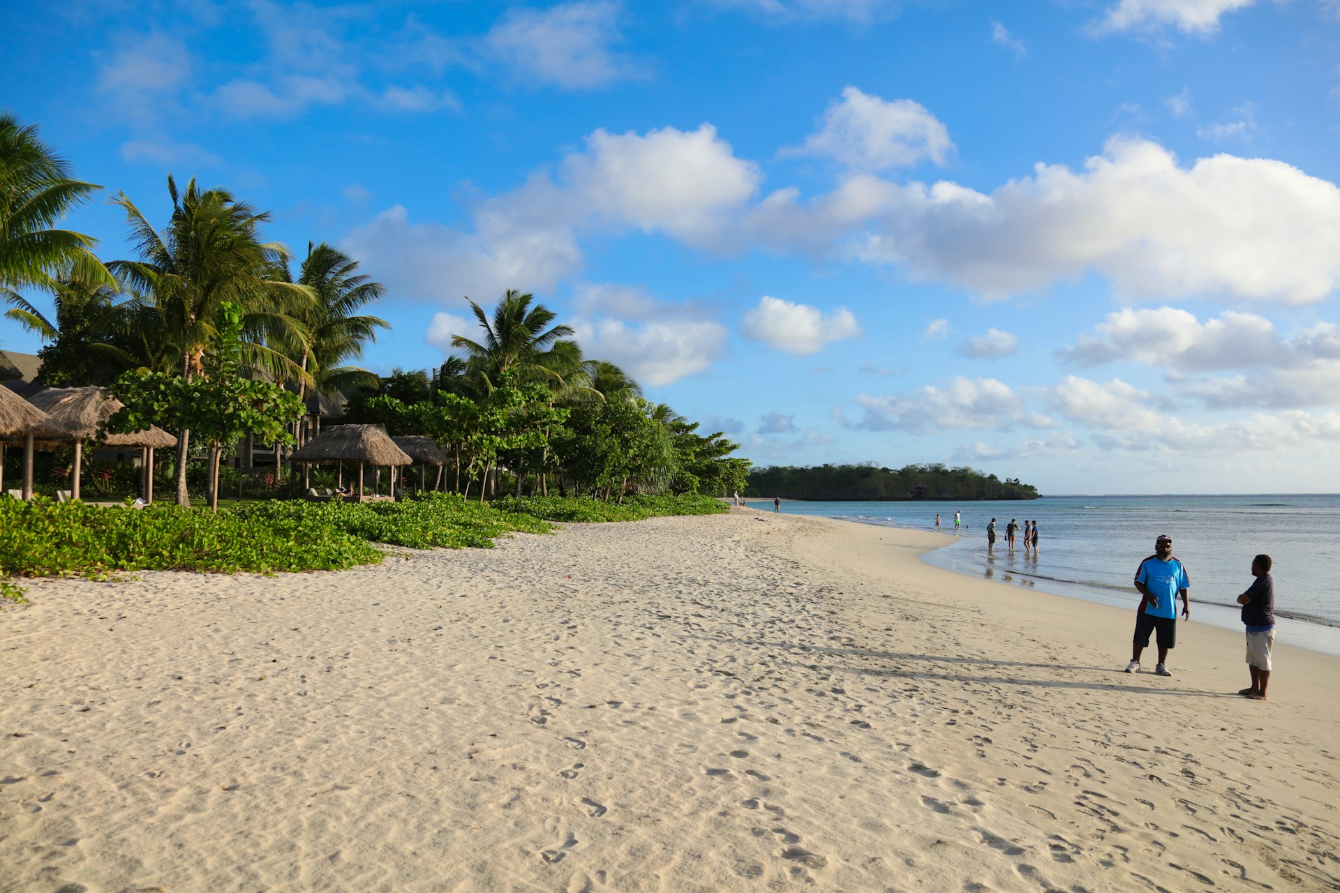 A wide section of sandy beach is backed by palm trees and lapped by blue water. A couple of rustic bungalows are visible amongst the palms behind the beach, while a few people paddle in the calm waters.