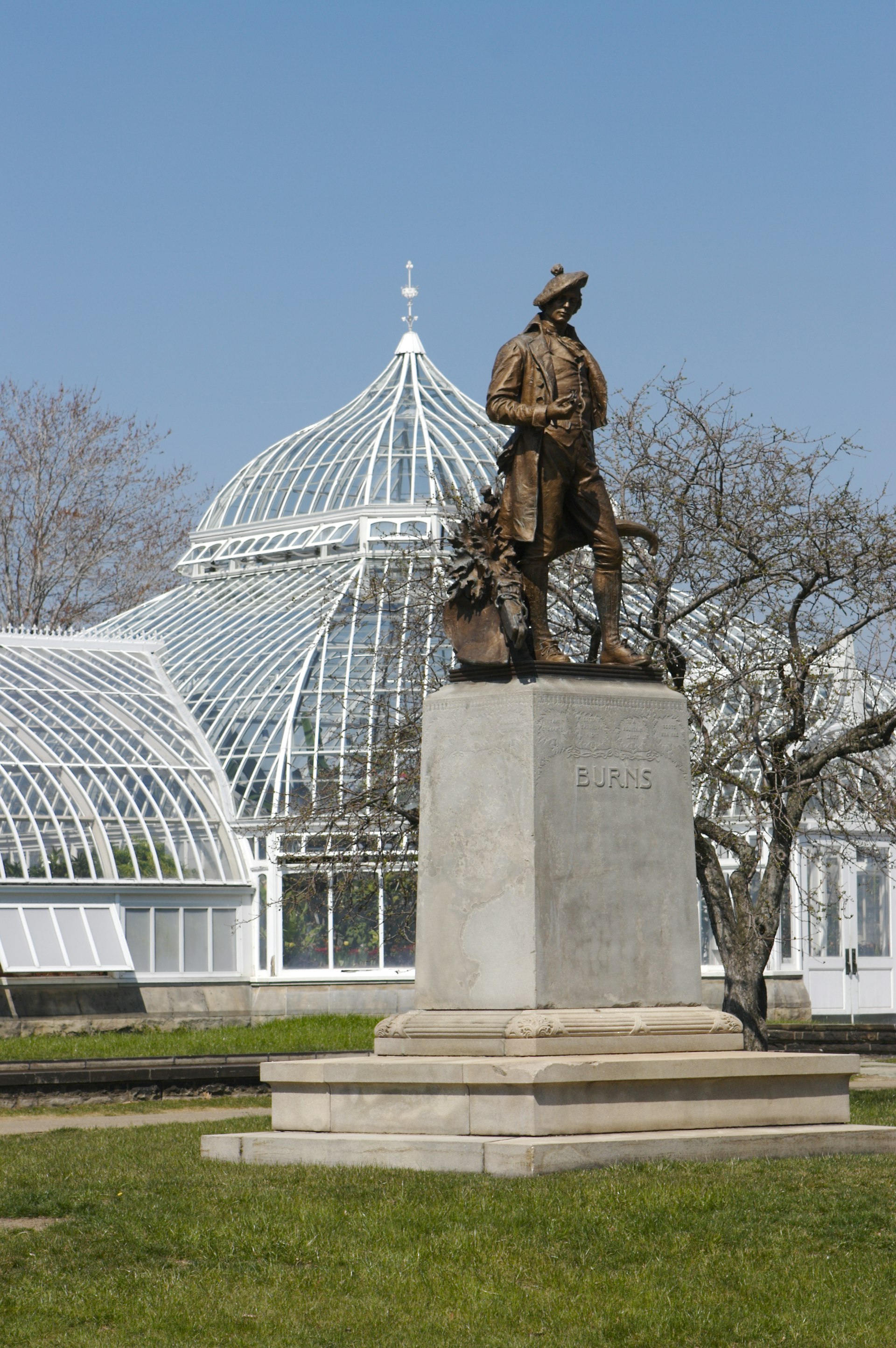 A bronze statue of a man wearing a hat, a large coat and knickerbockers stands in front of a glass green house at the Phipps Conservatory and Botanical Gardens in Pittsburgh.