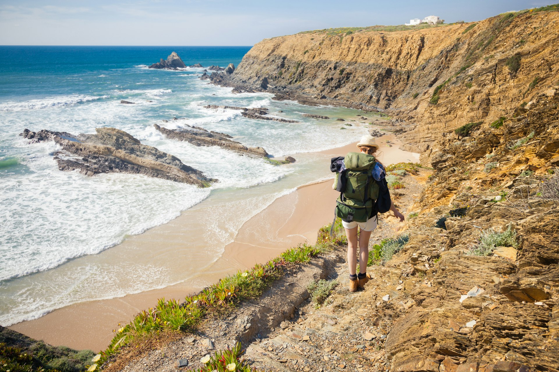 A female hiker with a large rucksack makes her way along the Rota Vicentina hiking trail in Portugal. Ahead of her is a wide sandy beach, which is being lapped by a calm sea.