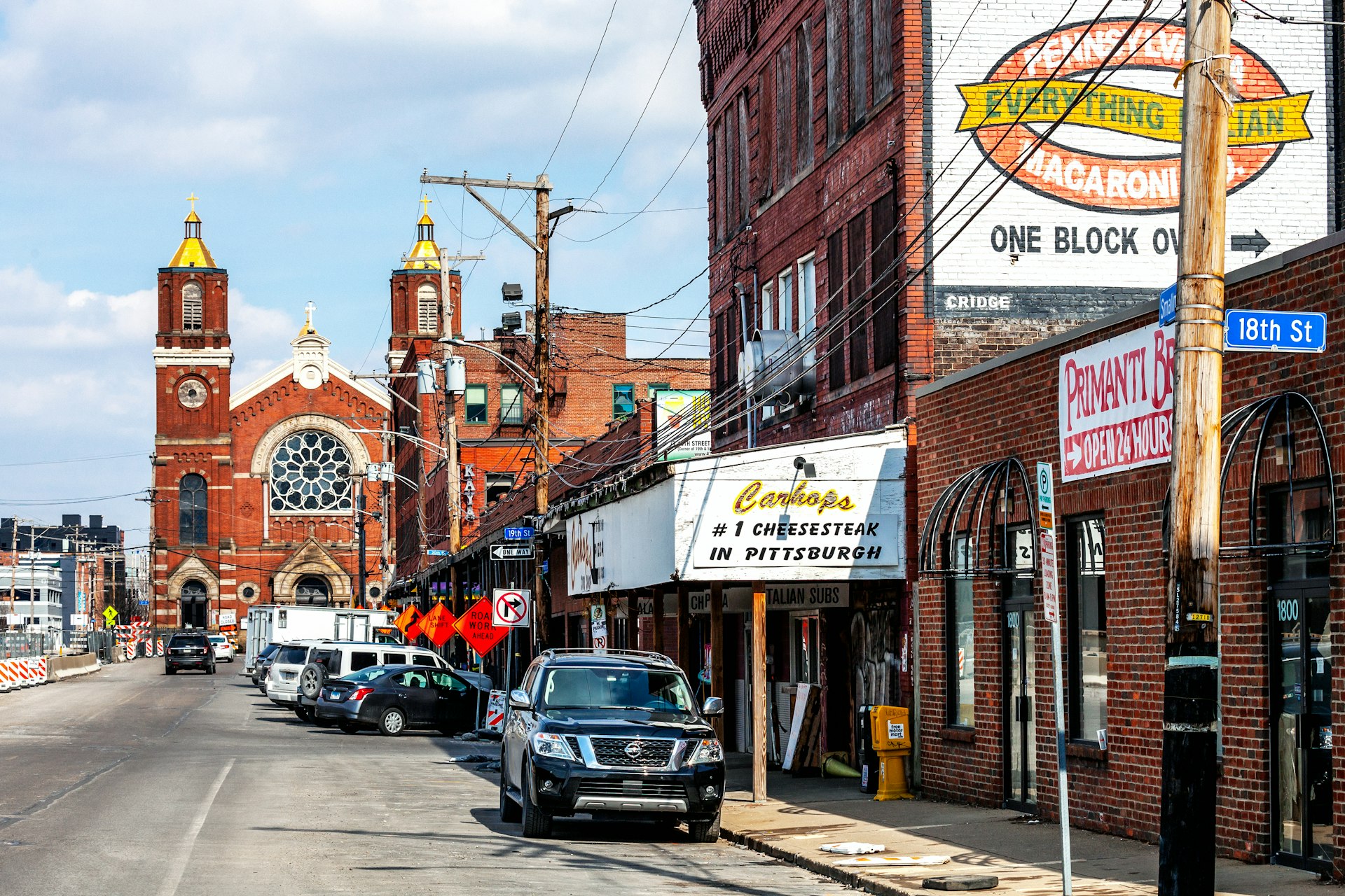 A row of brick businesses line the street in an area known as The Strip in Pittsburgh. Towards the end of the street is a light brown brick church. 