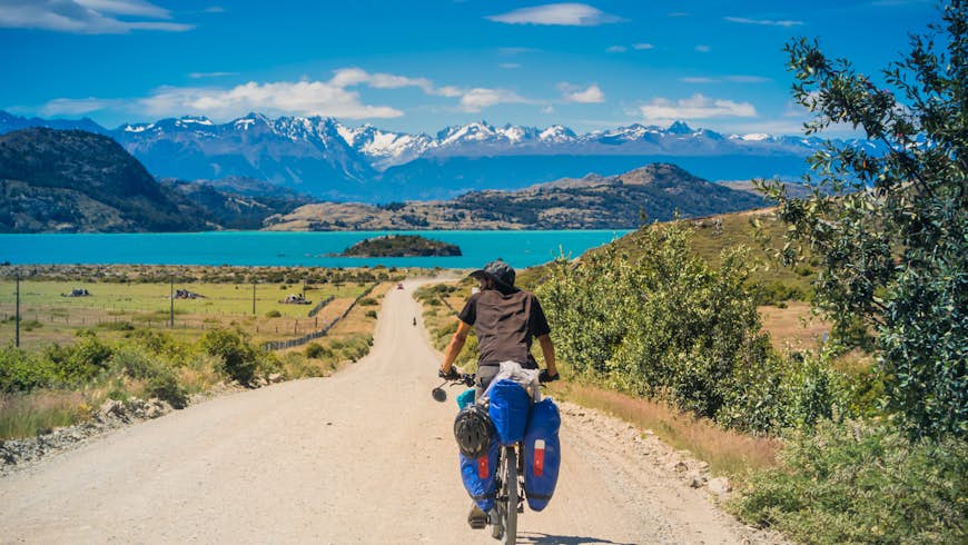 Ciclista masculino en camino de tierra en la Carretera Austral mientras se dirige al lago azul y las montañas grises.