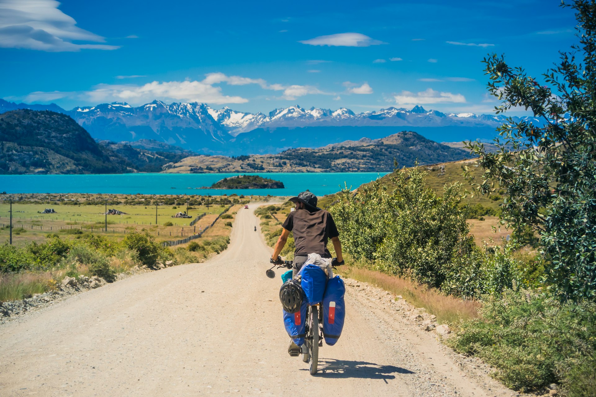 A male bicycle tourer on a dirt road of the Carretera Austral highway as it heads to a blue lake and gray mountains.