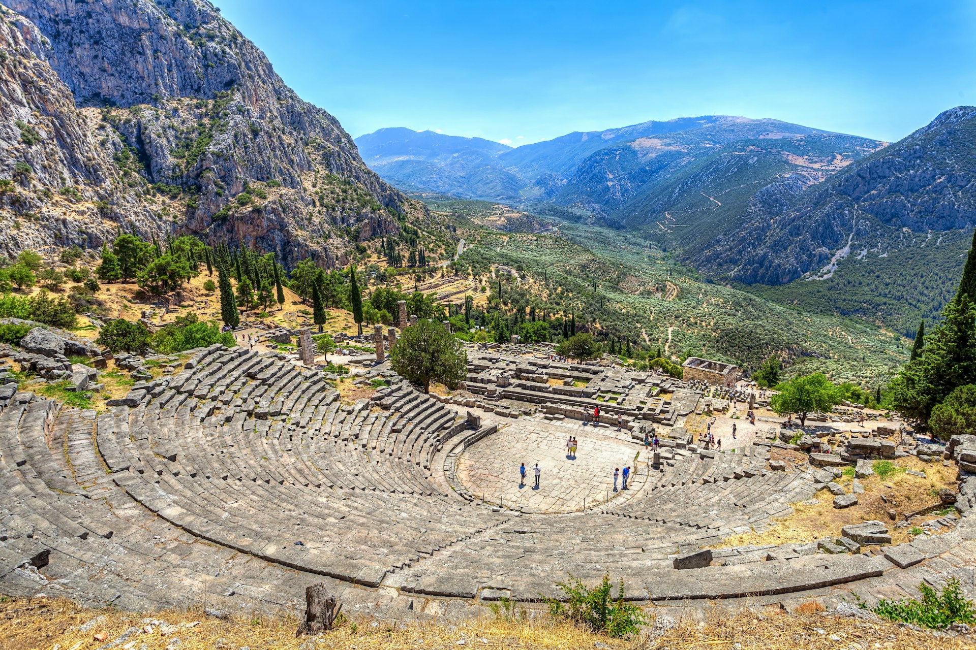 An ancient amphitheatre in Delphi, Greece. A few tourists stand in the large circular stone monument, while in the background vast green forests are visible.