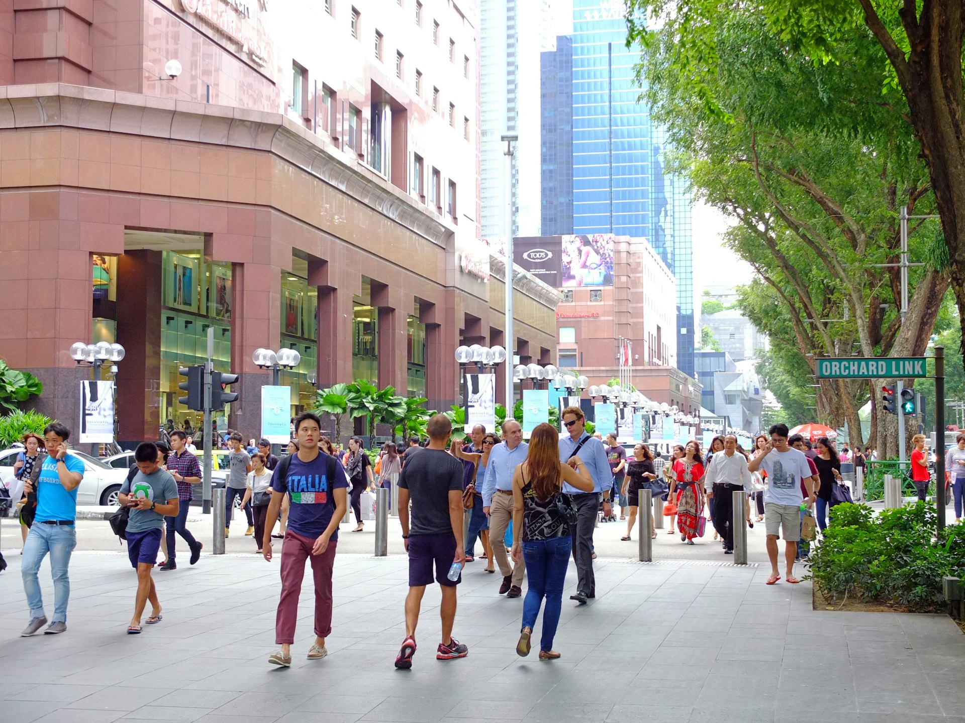 A crowd of people walk along Orchard Road on a sunny, blue-skied day in Singapore with a tall glass skyscraper rising up in the background.