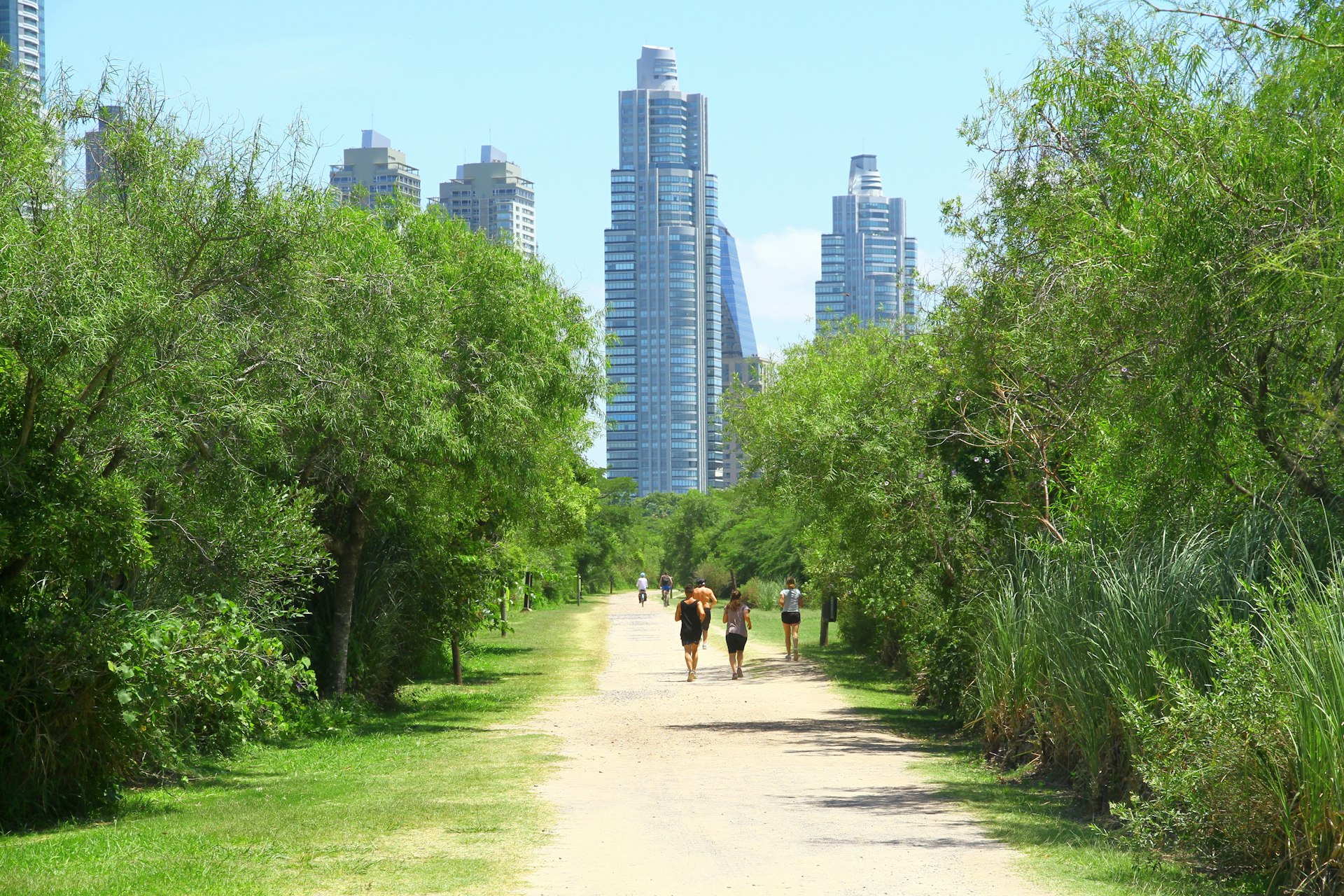 People walk and jog on a dirt path at Reserva Ecológica Costanera Sur with the skyline of Buenos Aires in the background