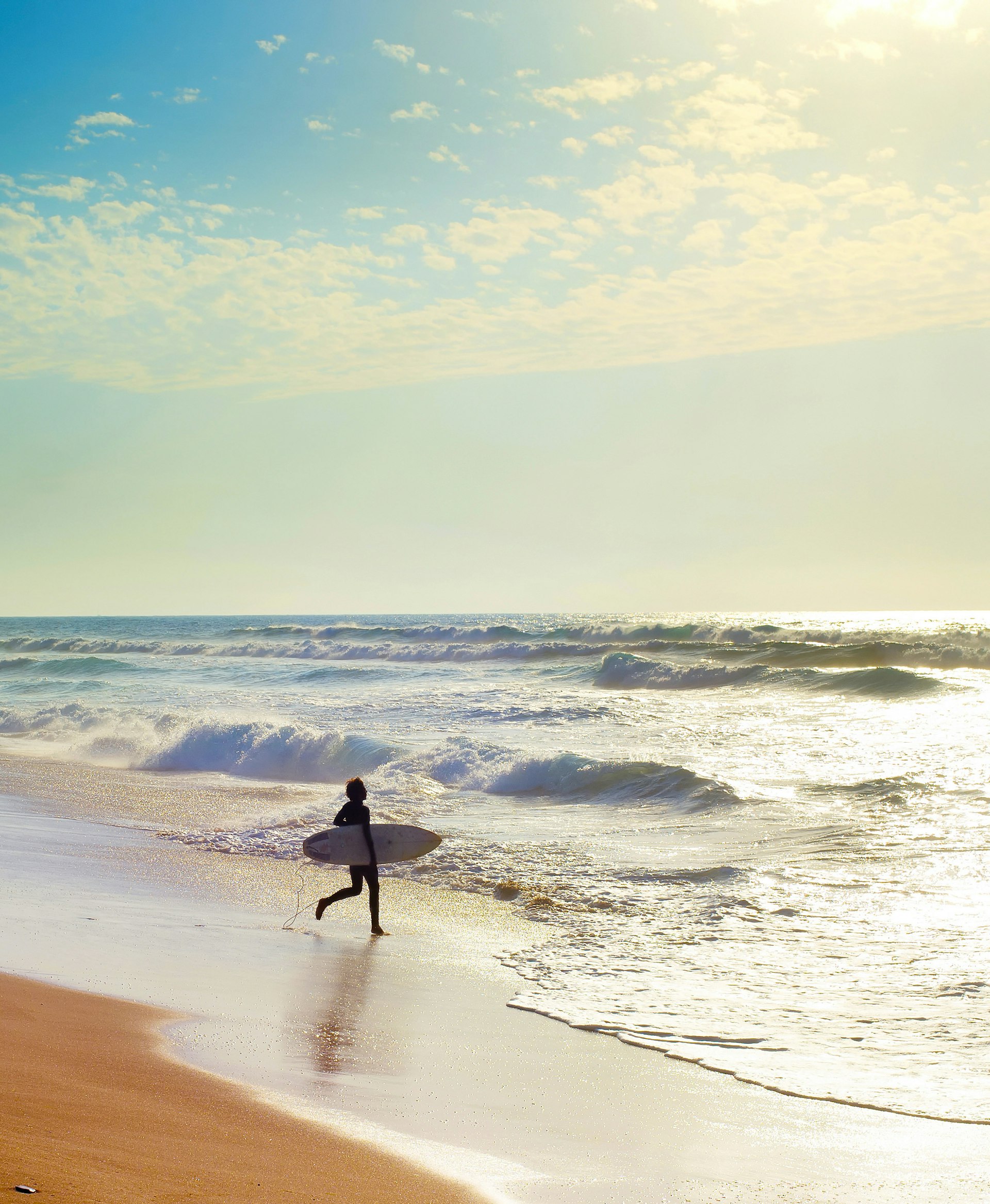 A surfer walks down the beach towards the sea in the blazing sunshine