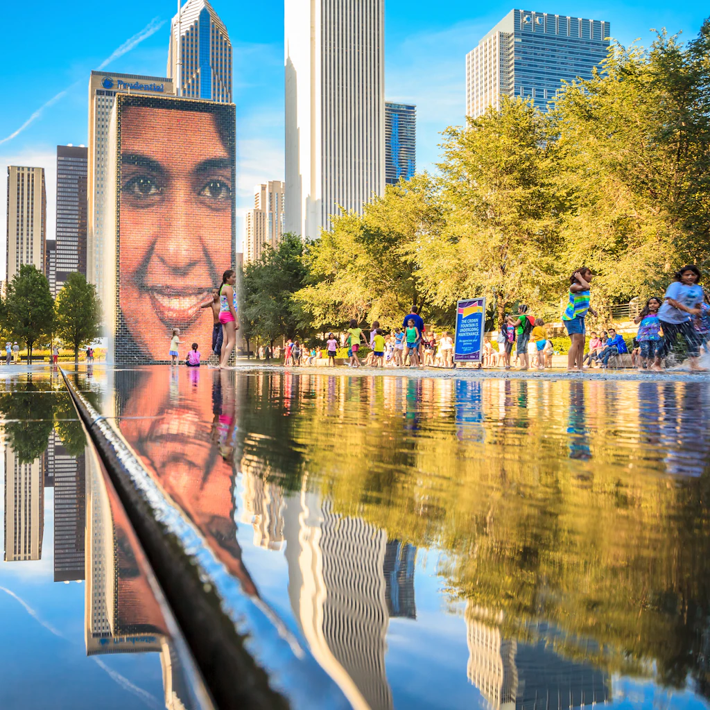 A large video screen with a smiling face stands above a water fountain in a park
