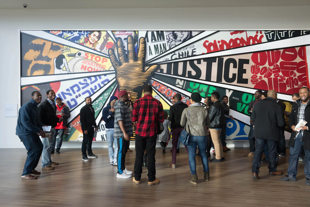 A group of people stand in front of a large painting with a black hand in the center and the word "justice" nearby