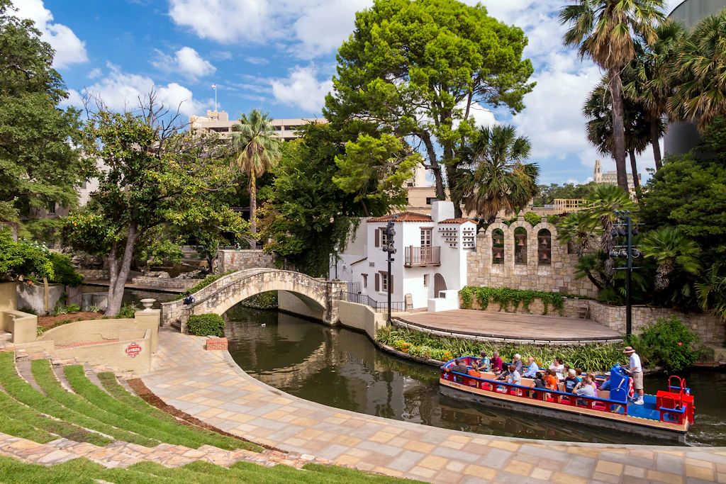 A small pleasure boat with around 10 passengers floats down a river towards a stone bridge