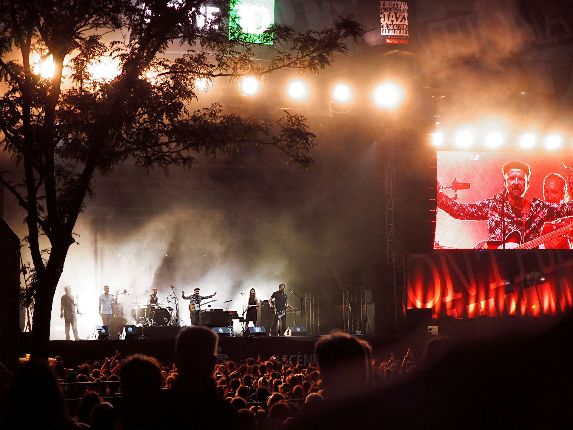 Shantel and Bocovina Orkestar perform a live concert during the Montreal Jazz Festival at night and are bathed in red light with a tree to the left of the stage