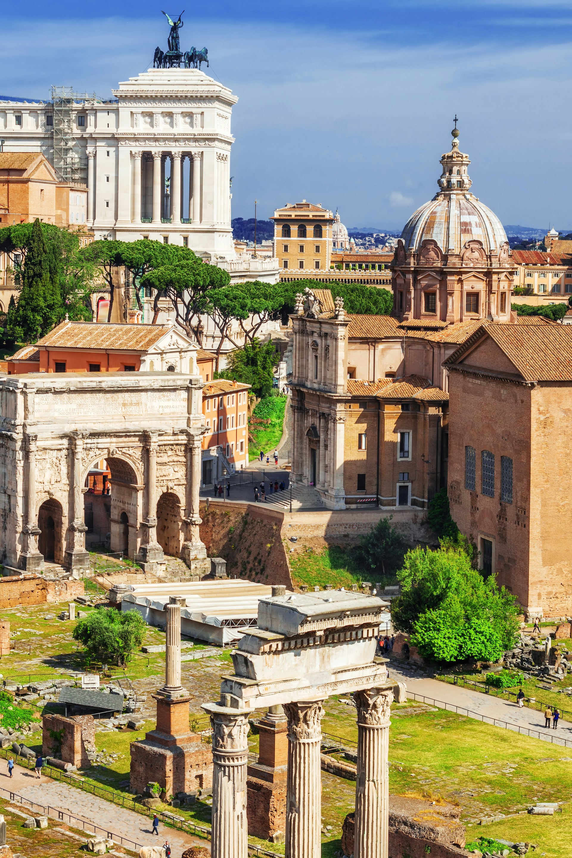 Ancient ruins viewed from above with a large building in the background with a bronze sculpture of horses on top