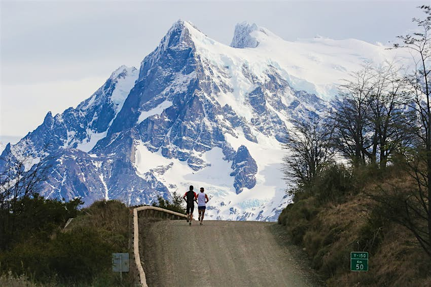 Los atletas que corren durante el Maratón Internacional Patagonia corren por la cresta de una carretera, con enormes montañas cubiertas de nieve al fondo.